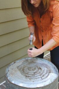 woman using awl and hammer to punch holes in to the bottom of a metal trash can